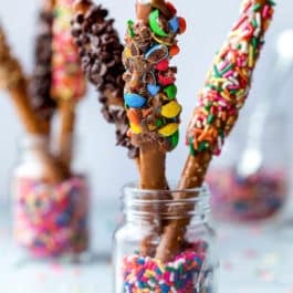 pretzel rods dipped in caramel and various toppings standing up in glass jars with sprinkles