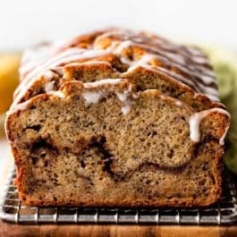 banana bread slice with cinnamon swirl and vanilla icing on top sitting on wire rack on cutting board.