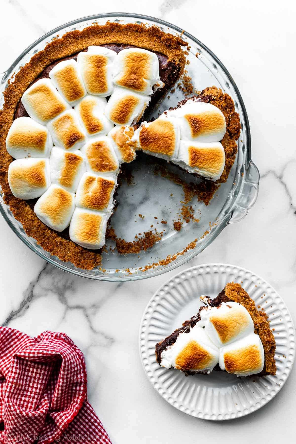 s'mores brownie pie in glass dish and slice shown on white plate.