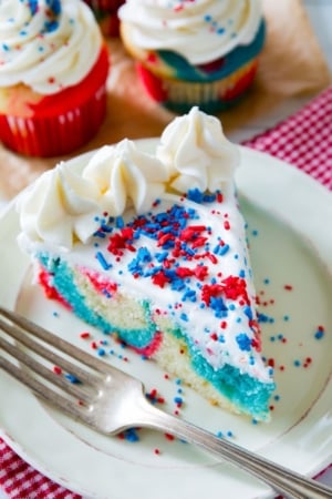 slice of red, white, and blue tie-dye cake topped with vanilla frosting and sprinkles on a white plate with a fork