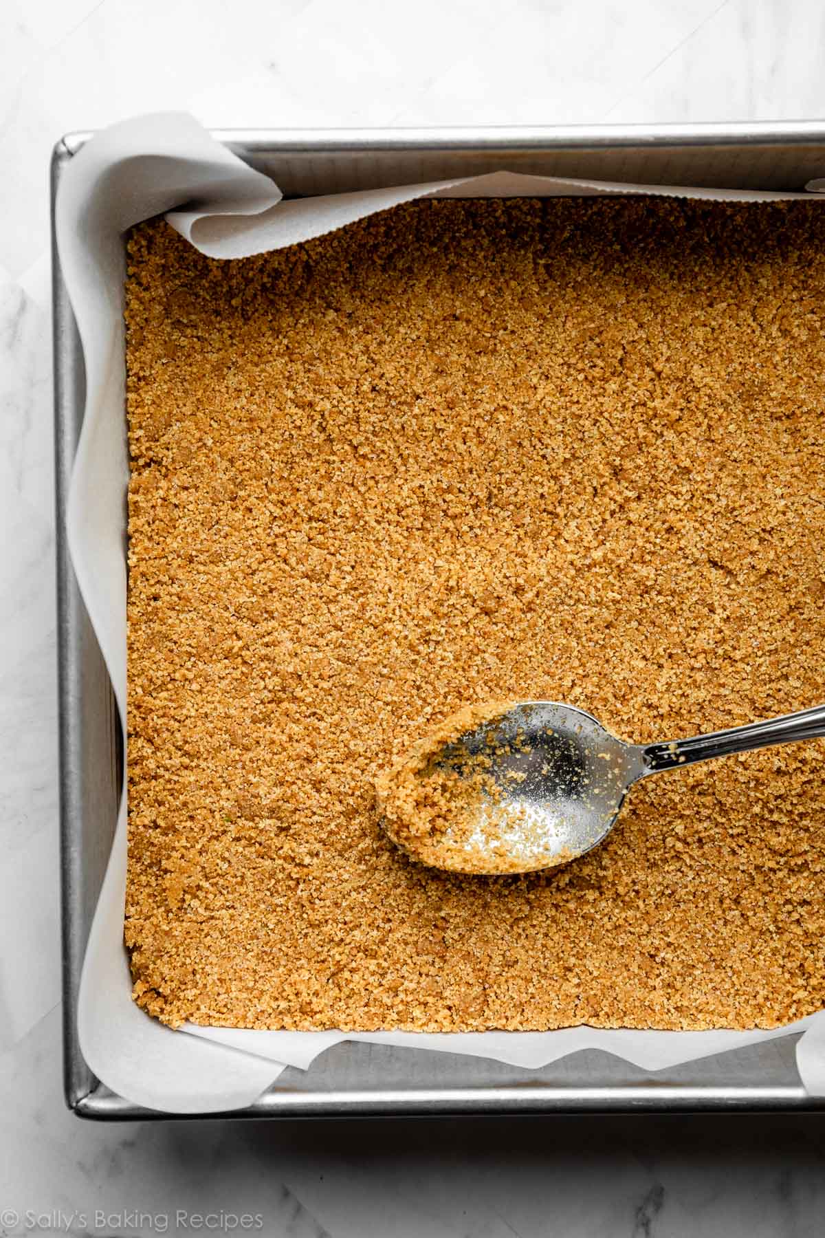 spoon flattening graham cracker crust in lined baking pan.