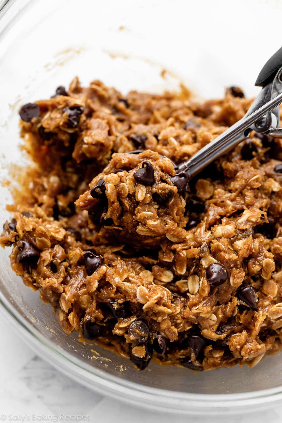 oatmeal cookie dough in glass bowl with cookie scoop scooping some out.