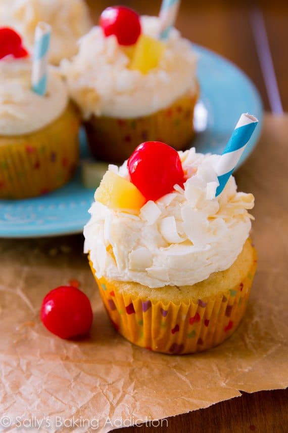 piña colada cupcakes topped with coconut frosting, shredded coconut, a cherry, slice of pineapple, and a straw