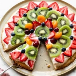 decorated sugar cookie fruit pizza on pizza pan with strawberries, kiwi, blueberries, grapes, and mandarin oranges on top.