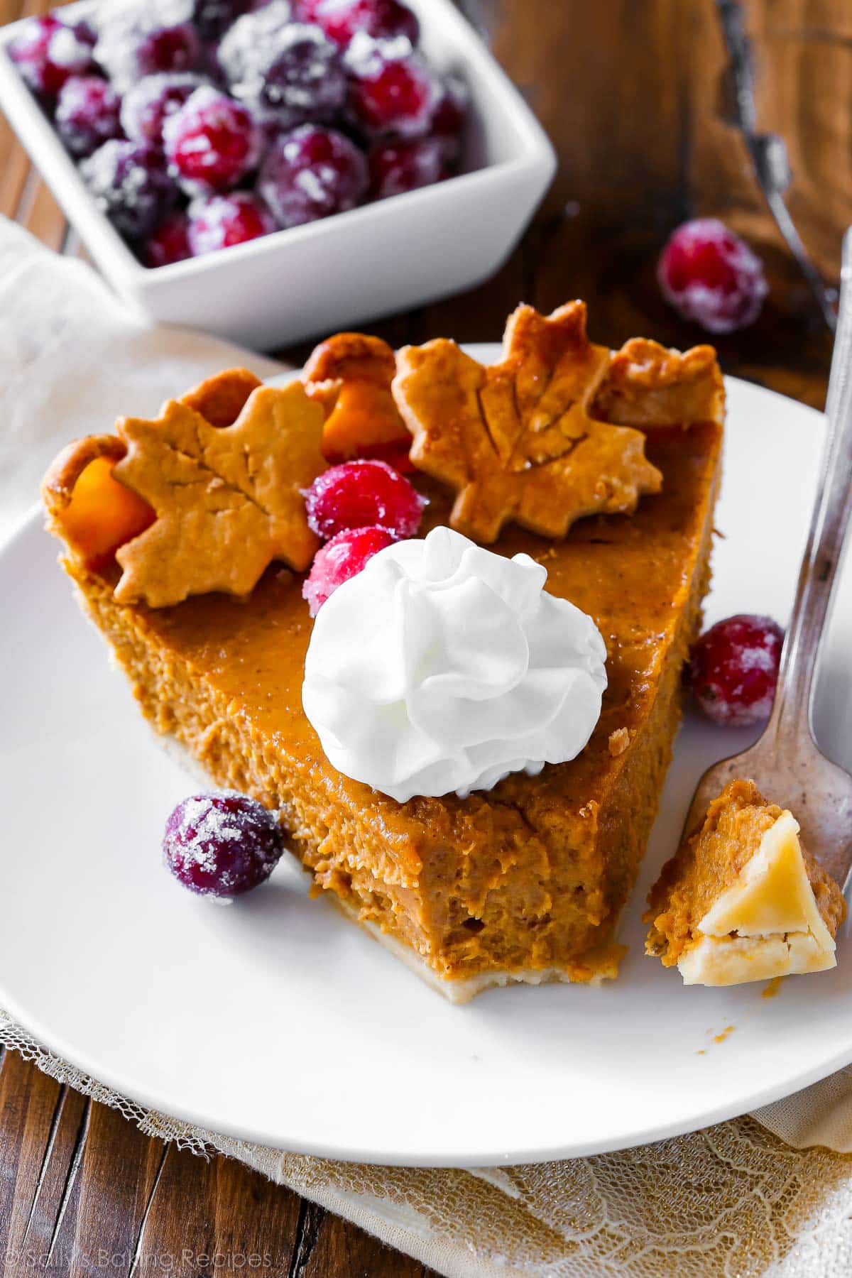 slice of pumpkin pie with forkful taken off with whipped cream, sugared cranberries, and pie crust leaf cut-outs on top.