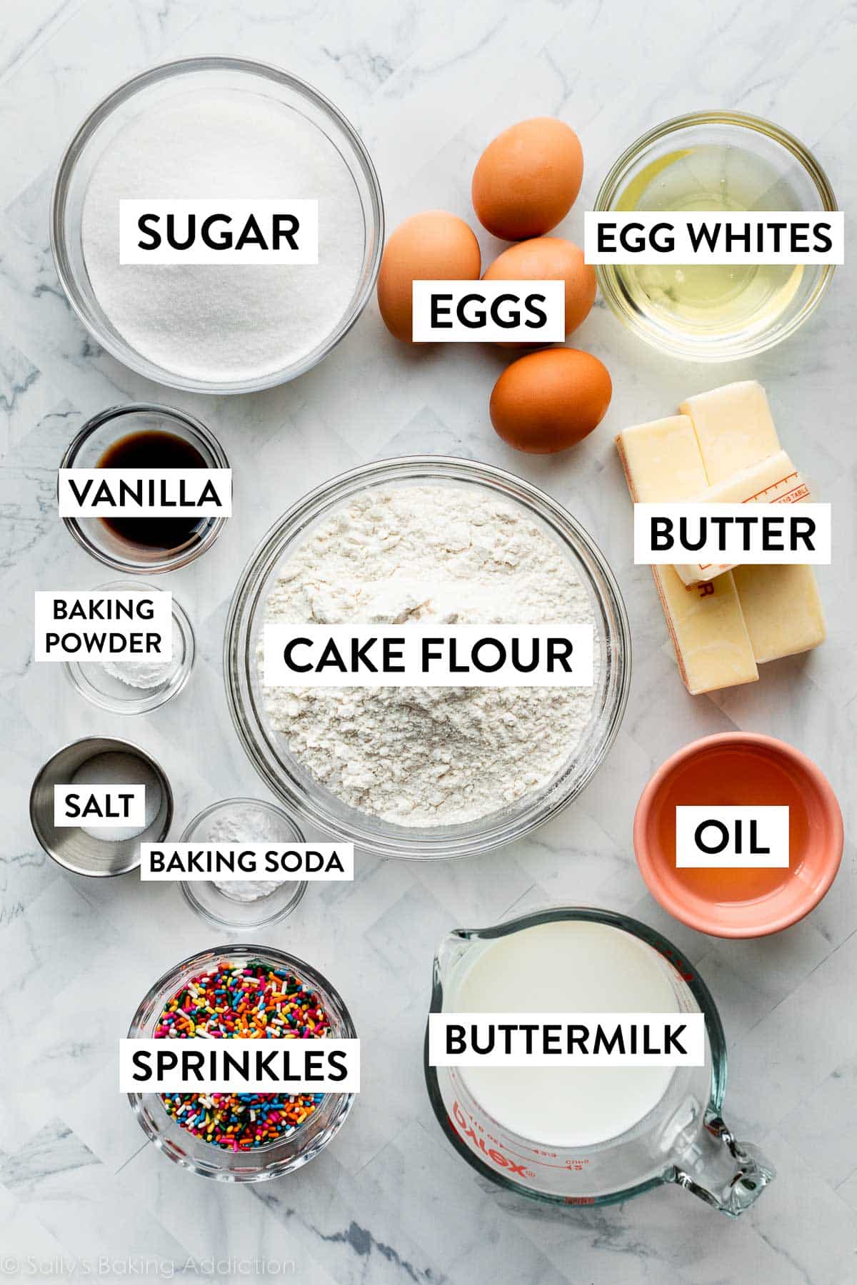 ingredients on marble counter including cake flour, butter, eggs, bowl of egg whites, oil, buttermilk, sprinkles, and more.