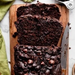 loaf of chocolate zucchini bread with chocolate chips cut with 2 slices on wooden cutting board.