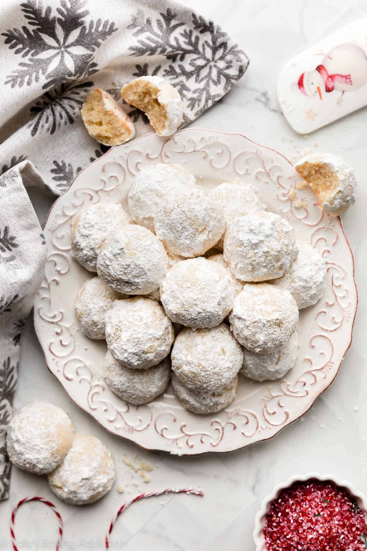 snowball cookies on white plate with snowflake linen and red sprinkles in bowl next to it.