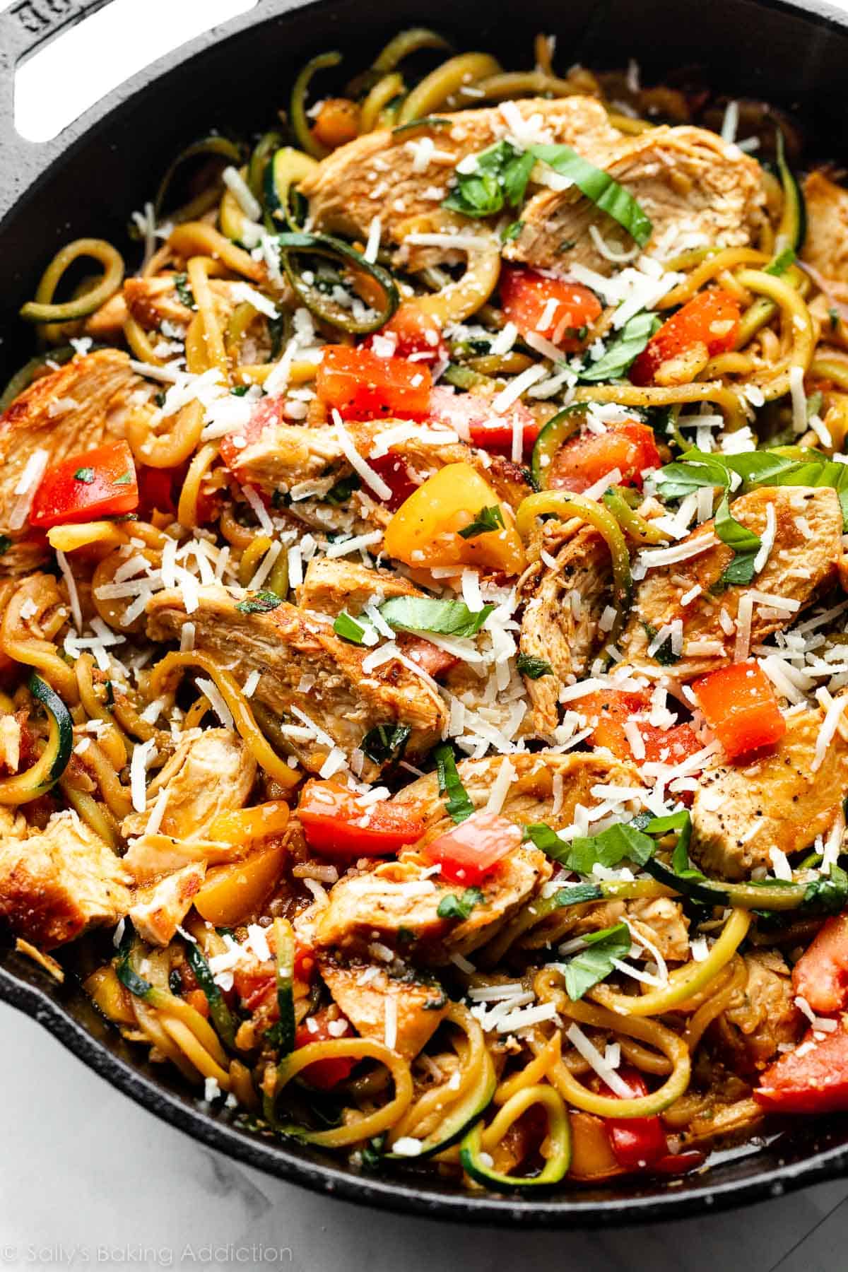 tomatoes and bruschetta mixture mixed with zucchini noodles, fresh basil and parmesan, and cooked chicken in a cast iron skillet.