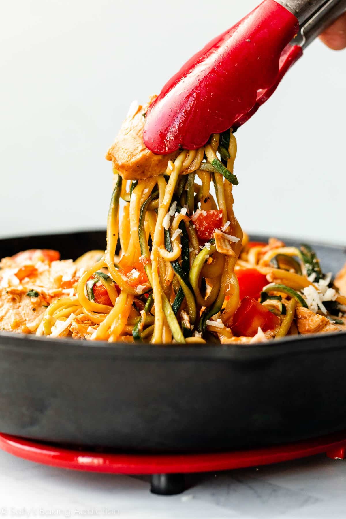 tongs pulling bruschetta zucchini noodles and chicken out of cast iron skillet.