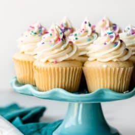 vanilla cupcakes on blue cake stand with pink, purple, and blue sprinkles on top.