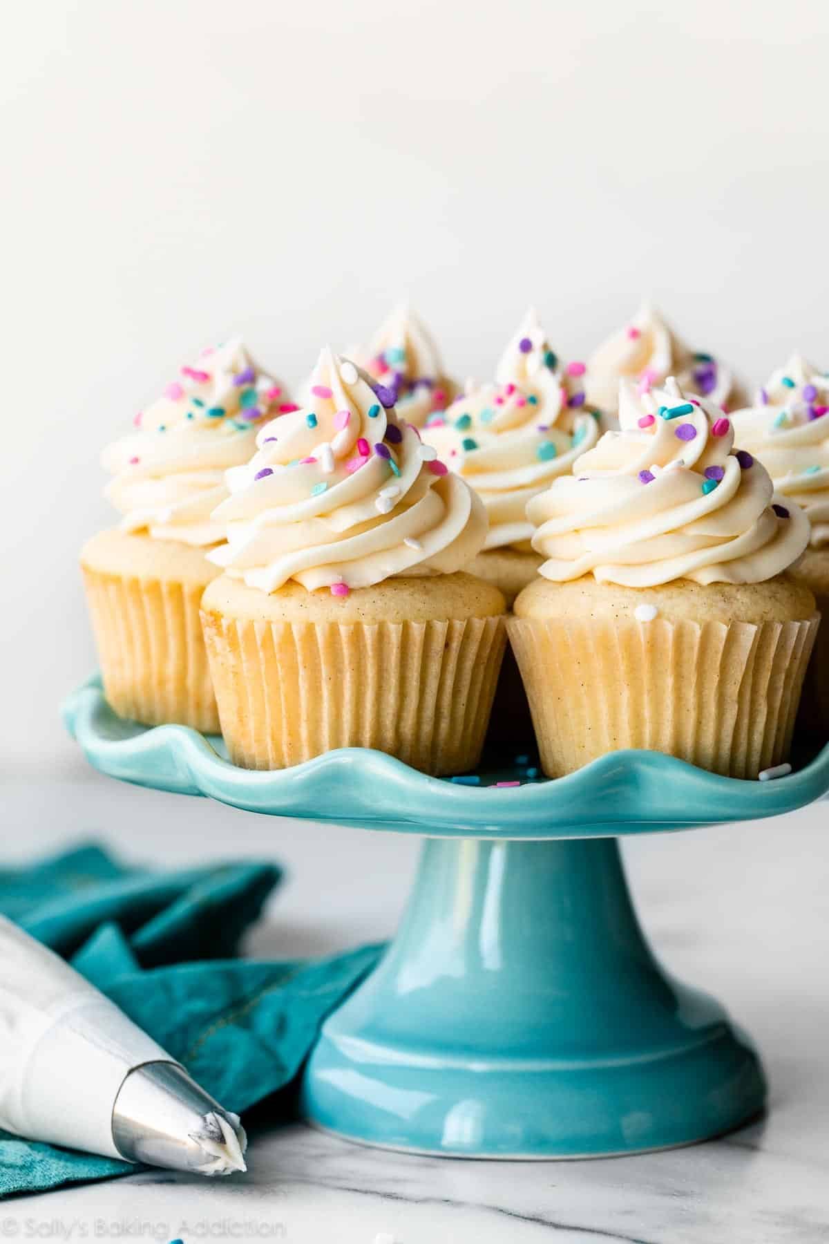 vanilla cupcakes on blue cake stand with pink, purple, and blue sprinkles on top.