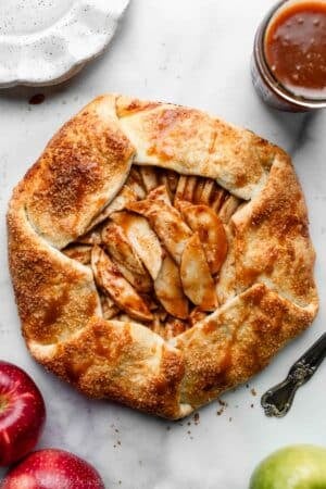 apple galette with caramel sauce on white parchment paper with apples and white plates surrounding it.