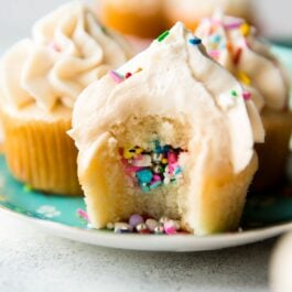 piñata cupcakes on a plate with a bite taken from one showing the sprinkles in the middle