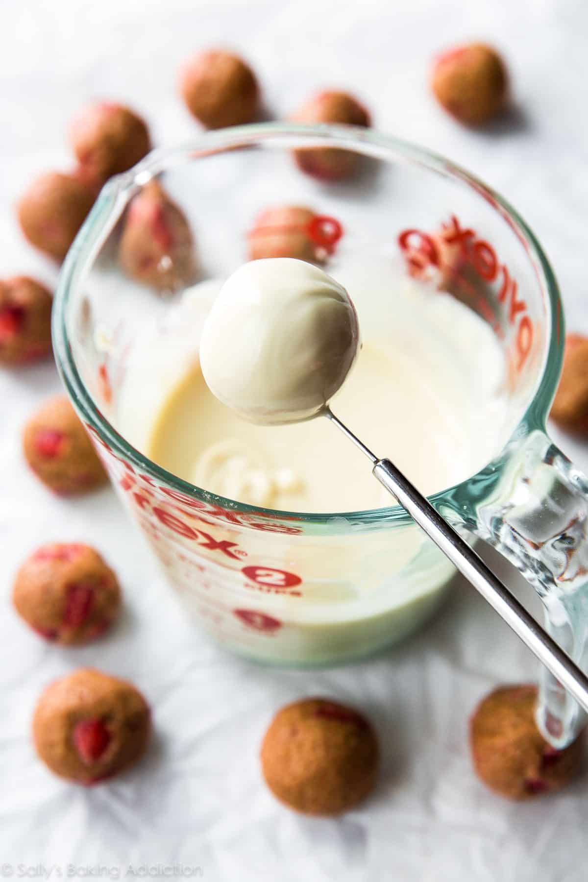 using a candy dipping tool to dip a truffle into white chocolate in a glass measuring cup