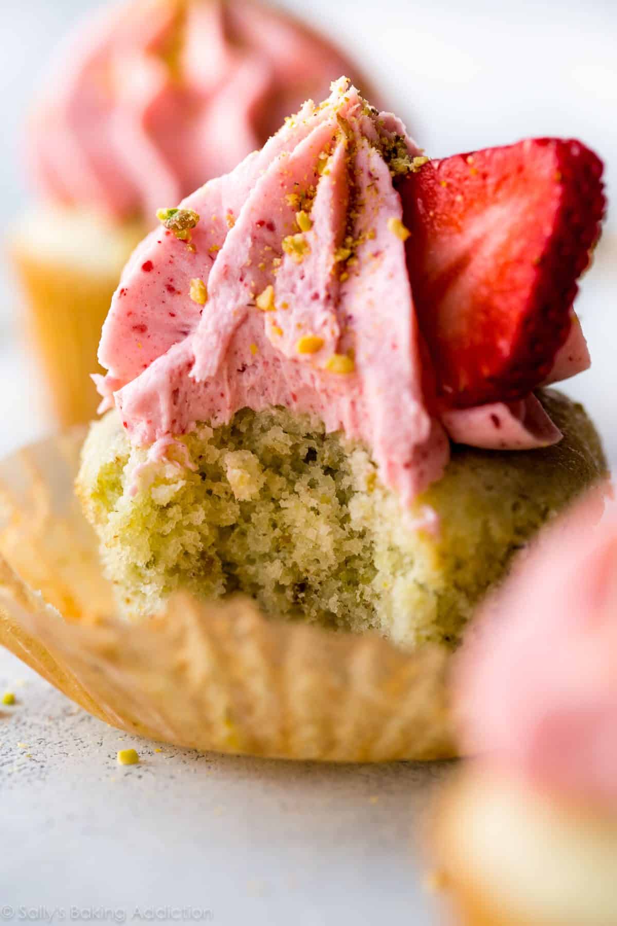 zoomed in image of a pistachio cupcake with a bite taken from it topped with strawberry frosting