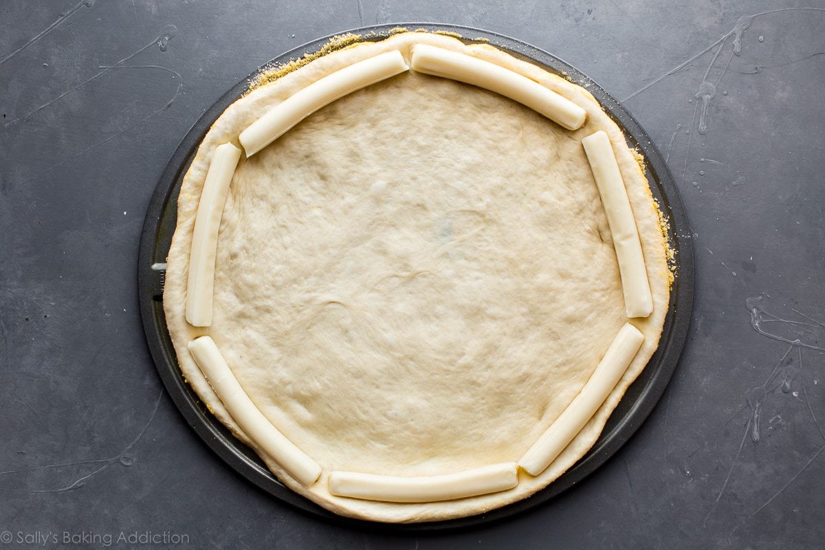 pizza dough rolled into a circle on a pizza pan with string cheese around the border of the dough