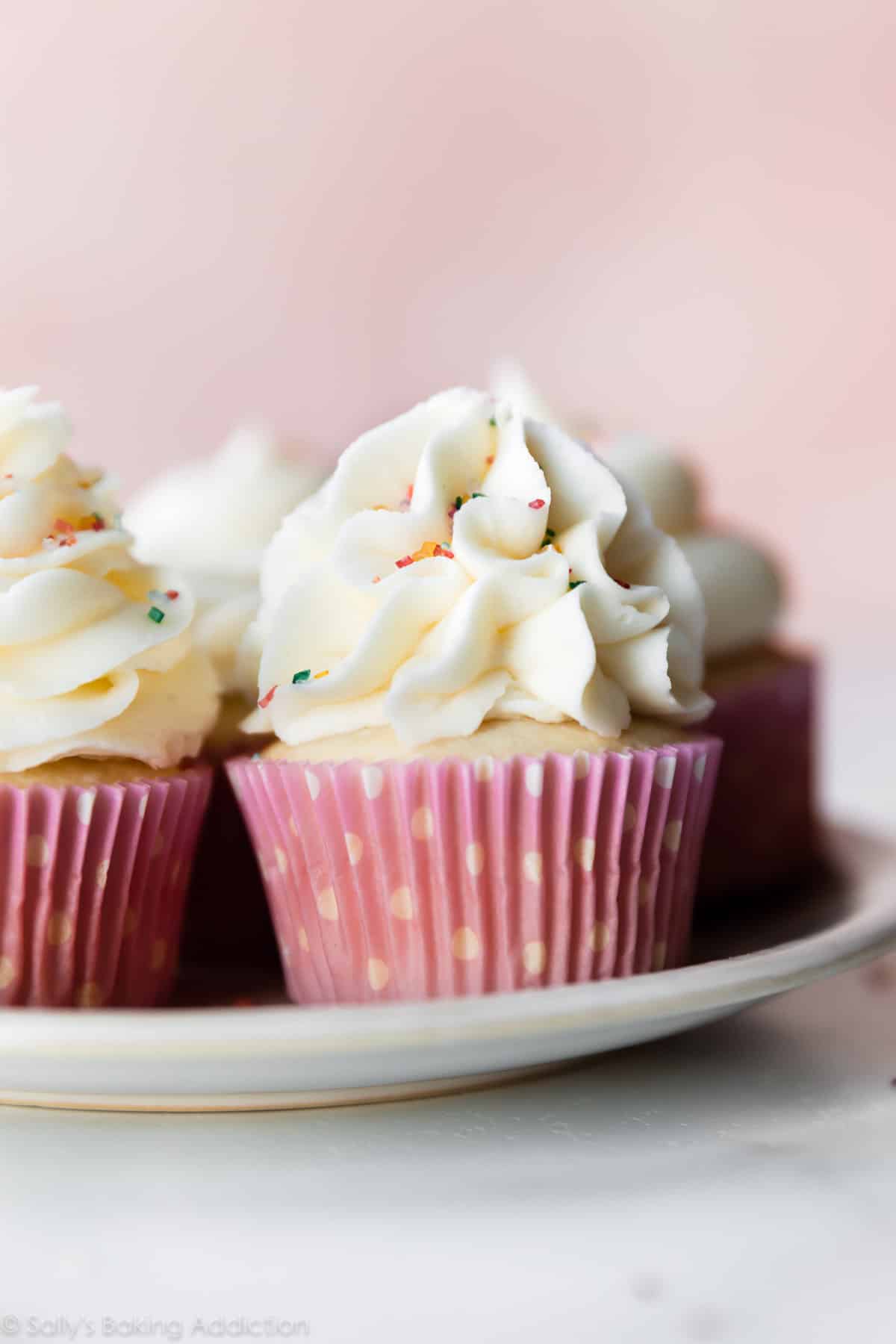 cupcakes on a plate topped with vanilla buttercream and sprinkles with pink polka dot cupcake wrappers