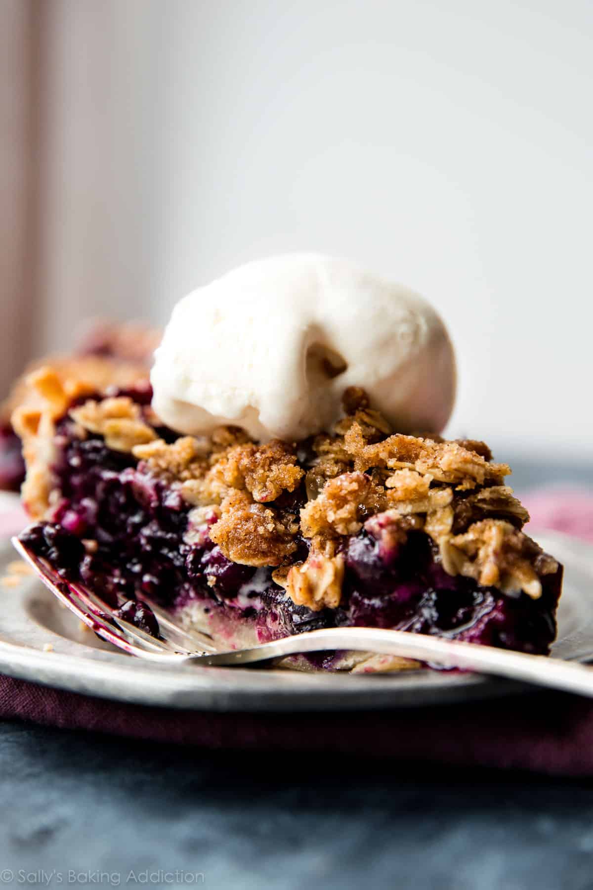 slice of blueberry crumble pie topped with a scoop of vanilla ice cream on a silver plate with a fork