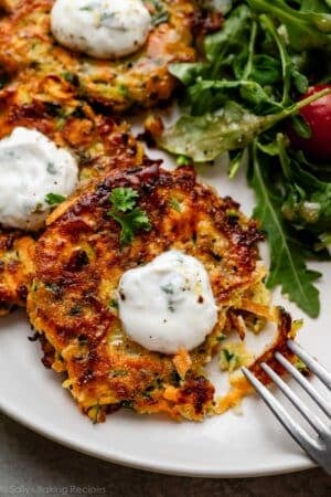 zucchini sweet potato fritter on place with yogurt sauce on top and green salad behind it.