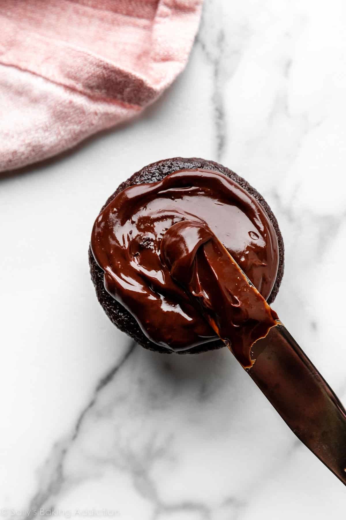 chocolate cupcake on marble counter with ganache being spread on top with icing spatula.