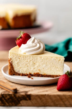 slice of cheesecake with whipped cream and strawberry on top on white plate on top of wood board.