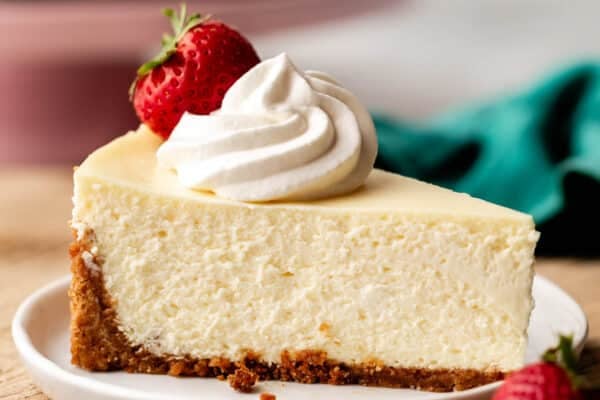 slice of cheesecake with whipped cream and strawberry on top on white plate on top of wood board.