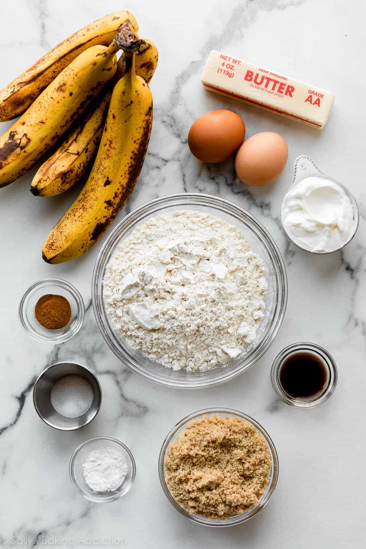 flour, brown sugar, vanilla, sour cream, butter, bananas, and other ingredients on marble counter.