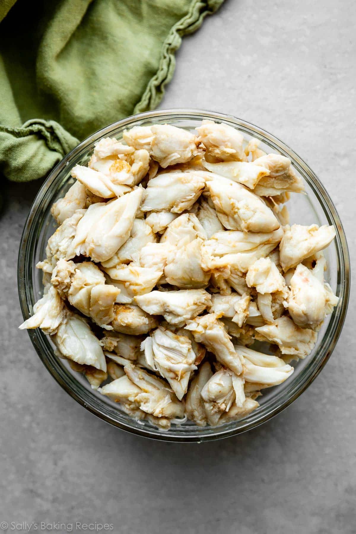 jumbo lump crab meat in glass bowl on gray backdrop with green linen in top left corner.