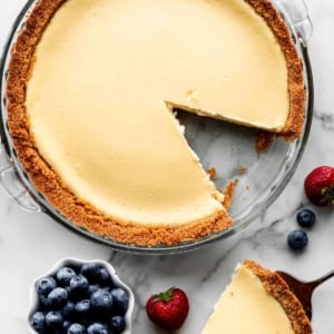 cheesecake pie in glass pan with slice taken out and blueberries and strawberries scattered next to the dish.