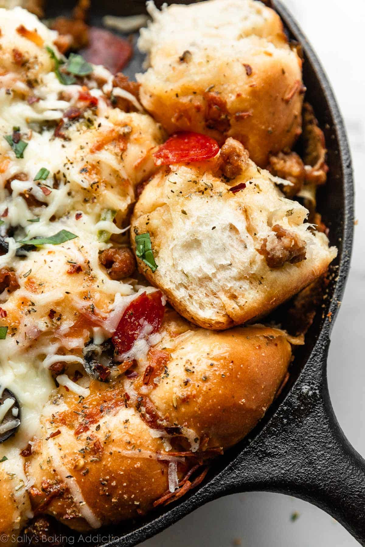 pizza pull apart rolls in cast iron skillet topped with cheese, pepperoni, olives, peppers, chopped basil, and sausage.