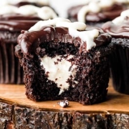 cream-filled chocolate cupcakes on wooden cake stand and 1 cupcake has bite taken out to reveal cream center.