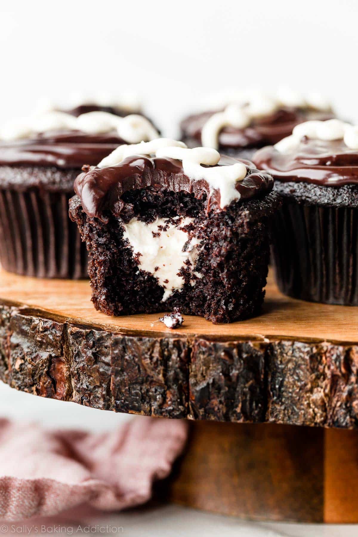 cream-filled chocolate cupcakes on wooden cake stand and 1 cupcake has bite taken out to reveal cream center.