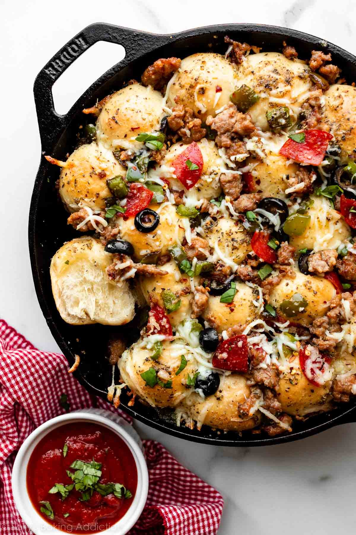 pizza pull apart rolls in cast iron skillet topped with cheese, pepperoni, olives, peppers, chopped basil, and sausage.