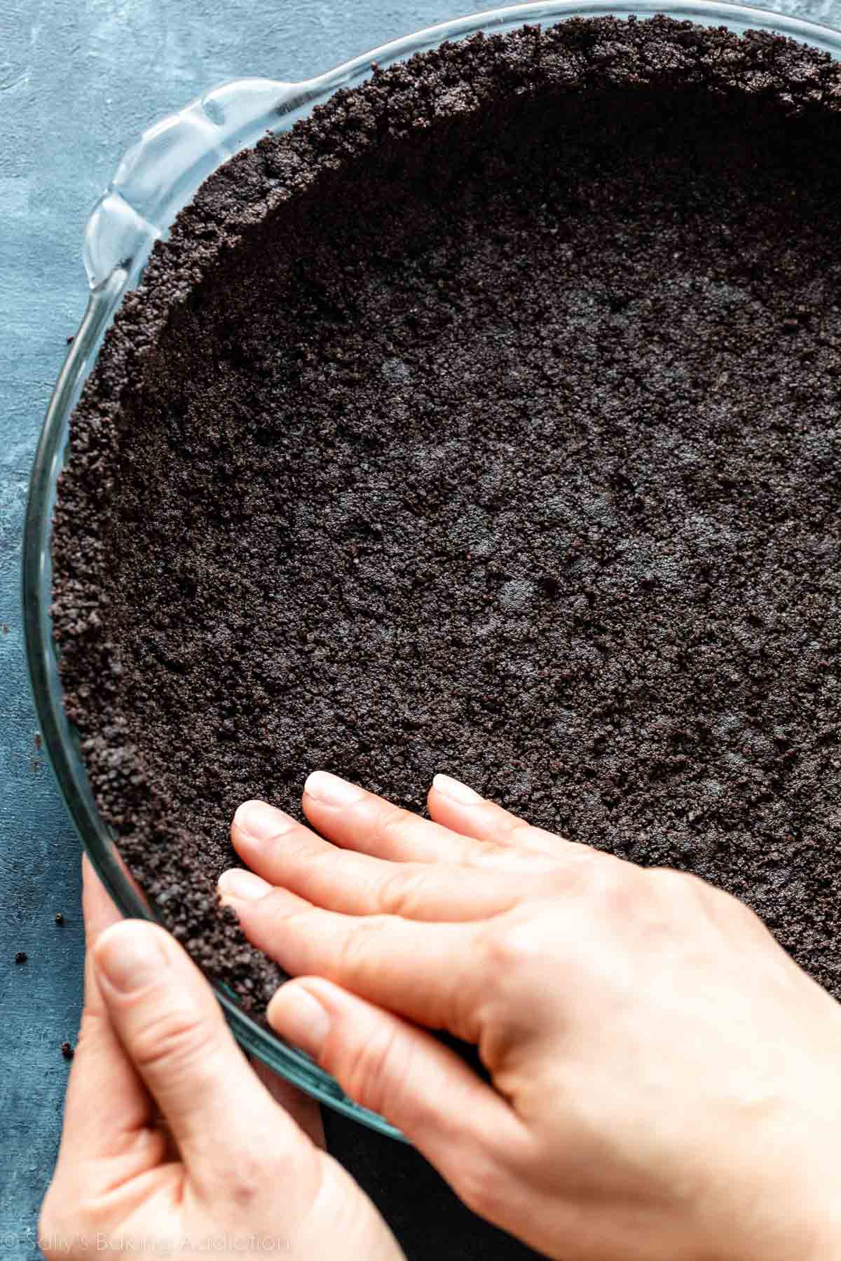 hands pressing Oreo cookie crust into pie dish on blue backdrop.