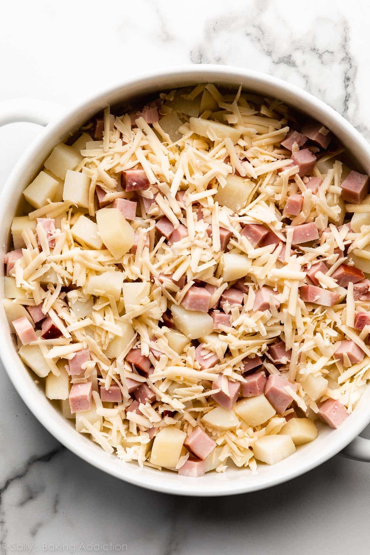 potatoes, ham, and shredded white cheese in round white ceramic baking dish.