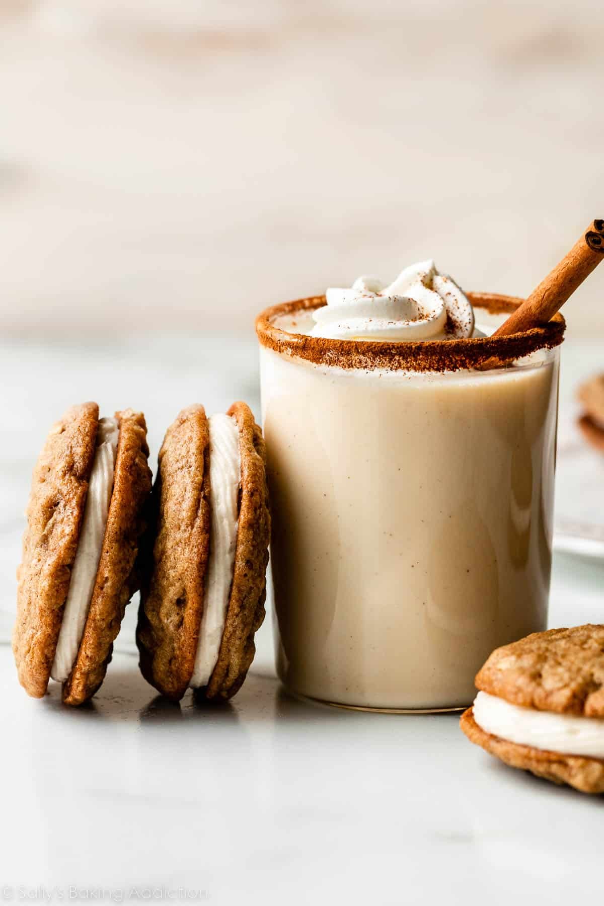 oatmeal cookies sandwiched with eggnog buttercream leaning on a cup of eggnog with whipped cream on top.