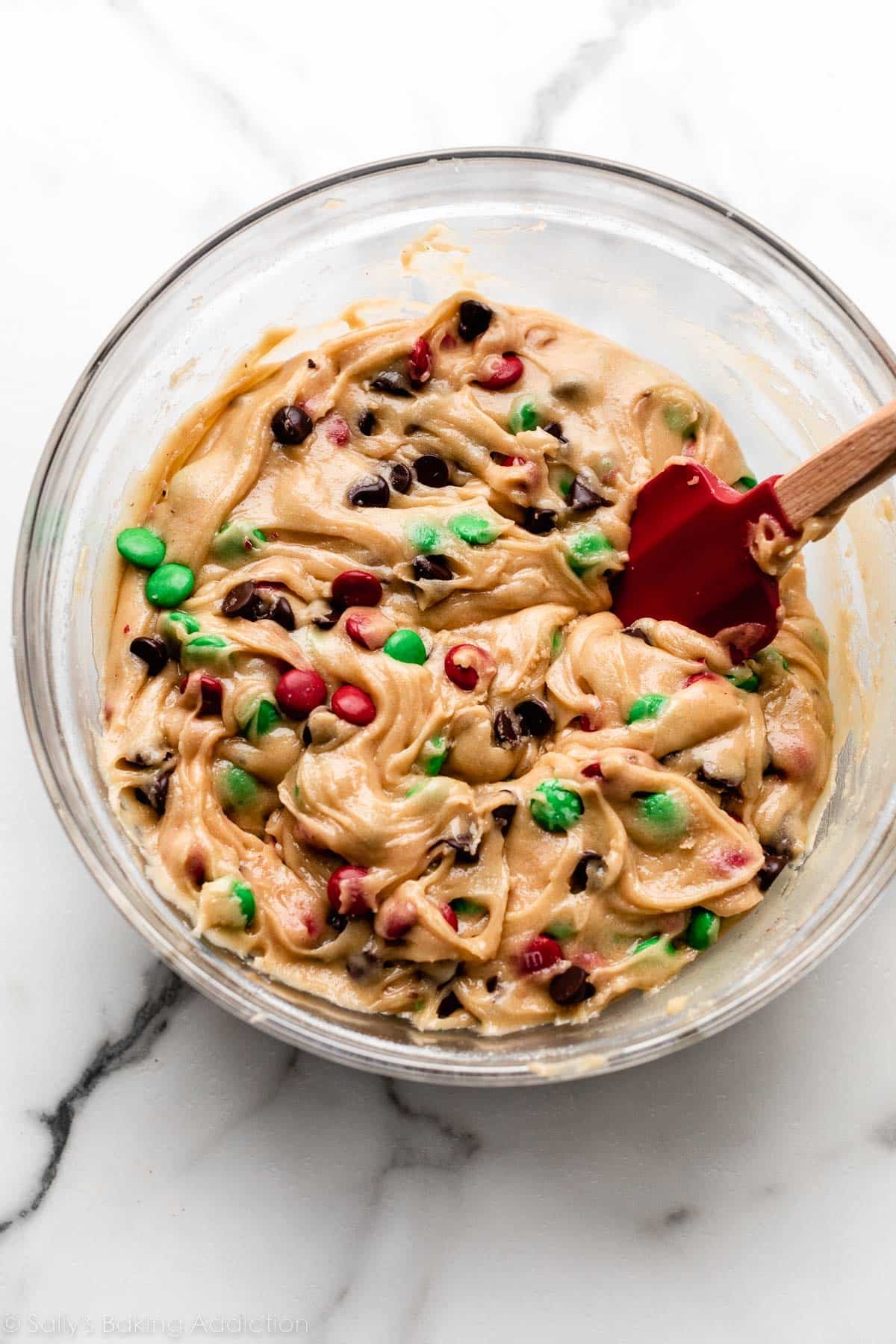 chocolate chip cookie dough in glass bowl with Christmas-colored M&Ms and red spatula.