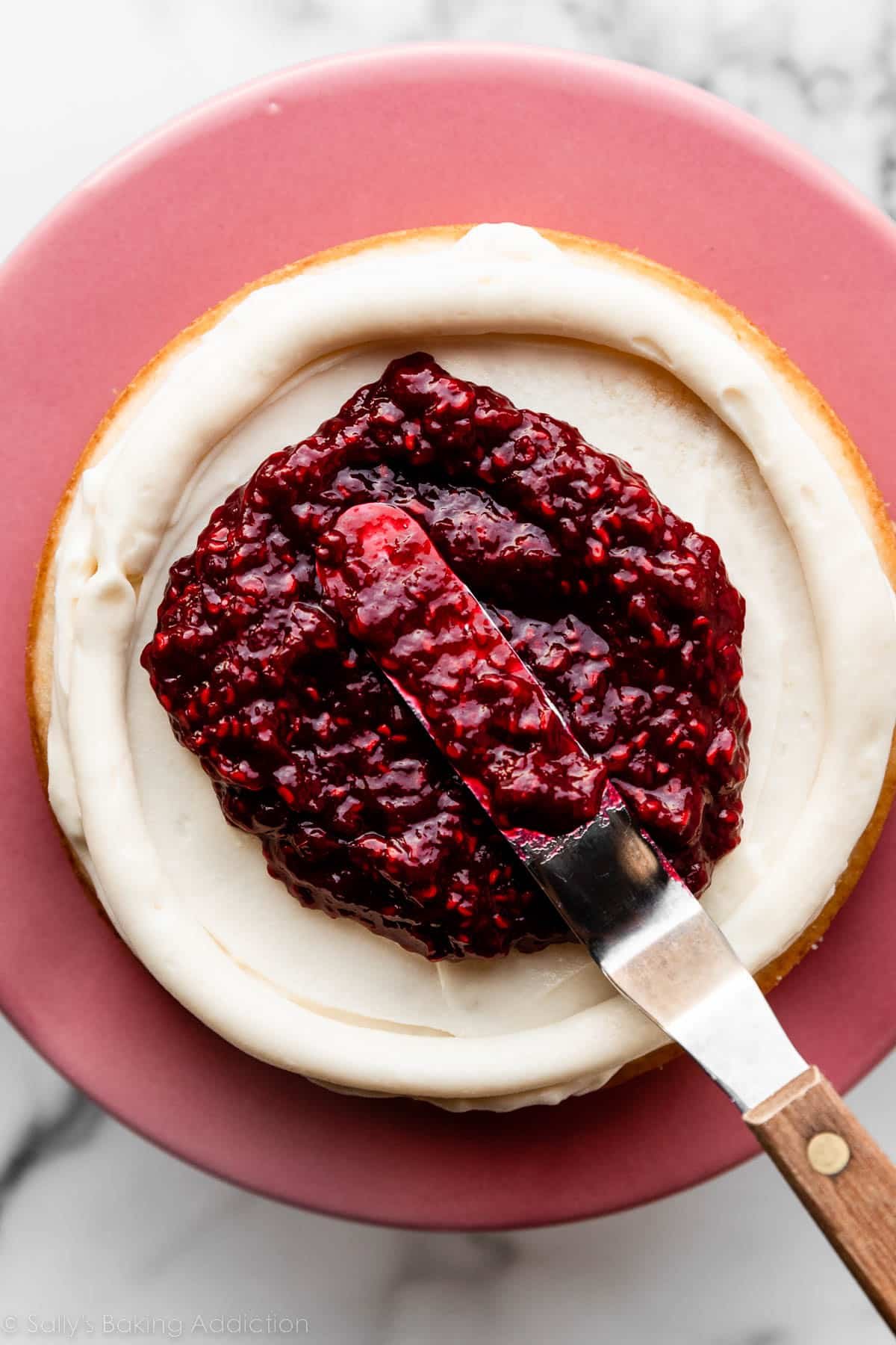 small offset spatula shown spreading raspberry filling on layer of cake with white frosting underneath and around it.