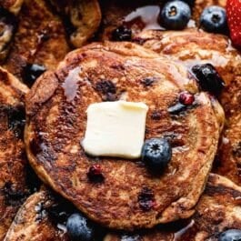 close-up of whole wheat blueberry pancakes with maple syrup, butter, and fresh blueberries on top.