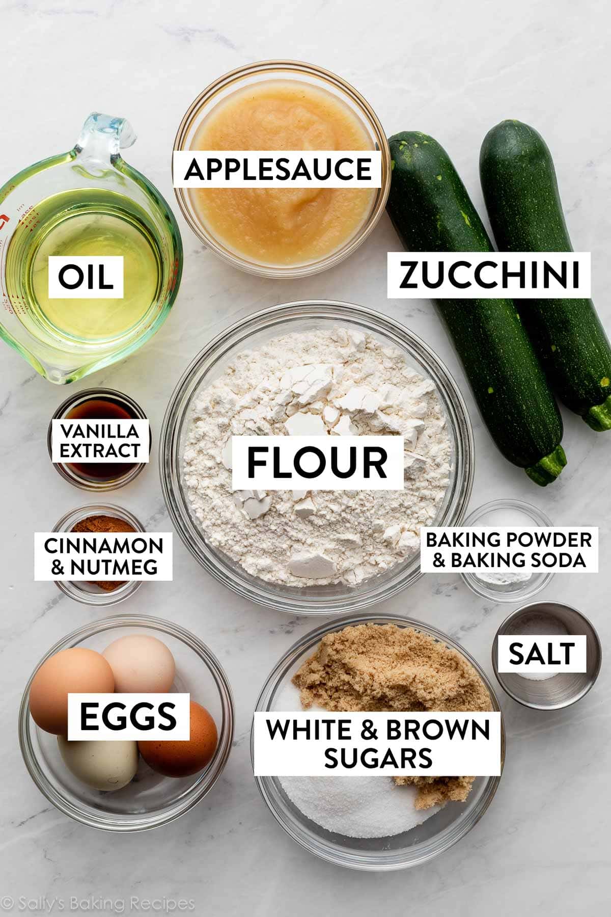 ingredients on marble counter including applesauce, flour, sugar, eggs, oil, cinnamon and nutmeg, and vanilla extract.