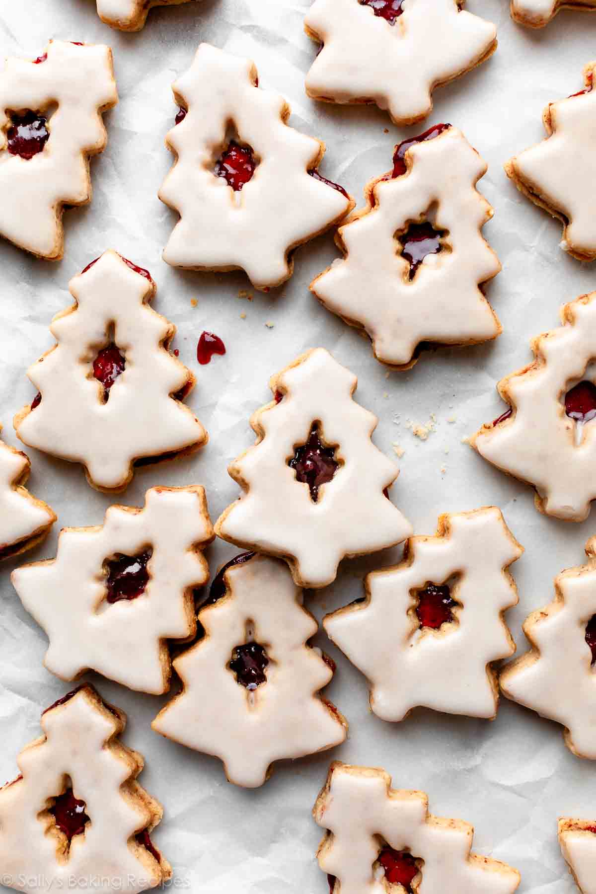 Christmas-tree shaped cherry almond linzer cookies with icing on white parchment paper.