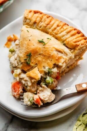 slice of double crust chicken pot pie with golden flaky pie crust on white plate with fork that has a wooden handle.