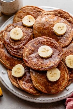 big plate of whole wheat banana pancakes with maple syrup and banana slices.